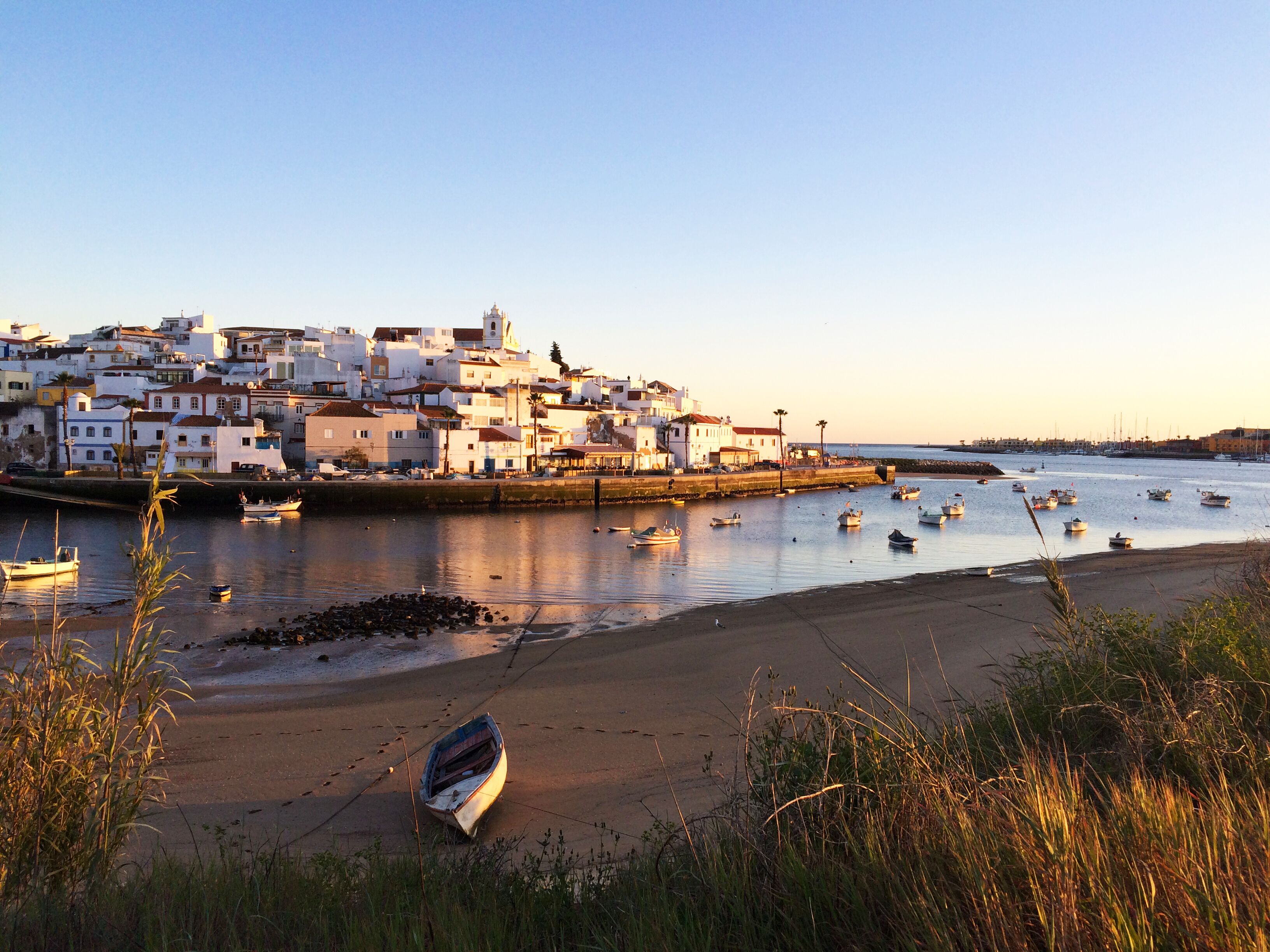 Un pueblo costero con edificios a lo largo del paseo marítimo, barcos en el agua y una playa con una sola embarcación en la arena.