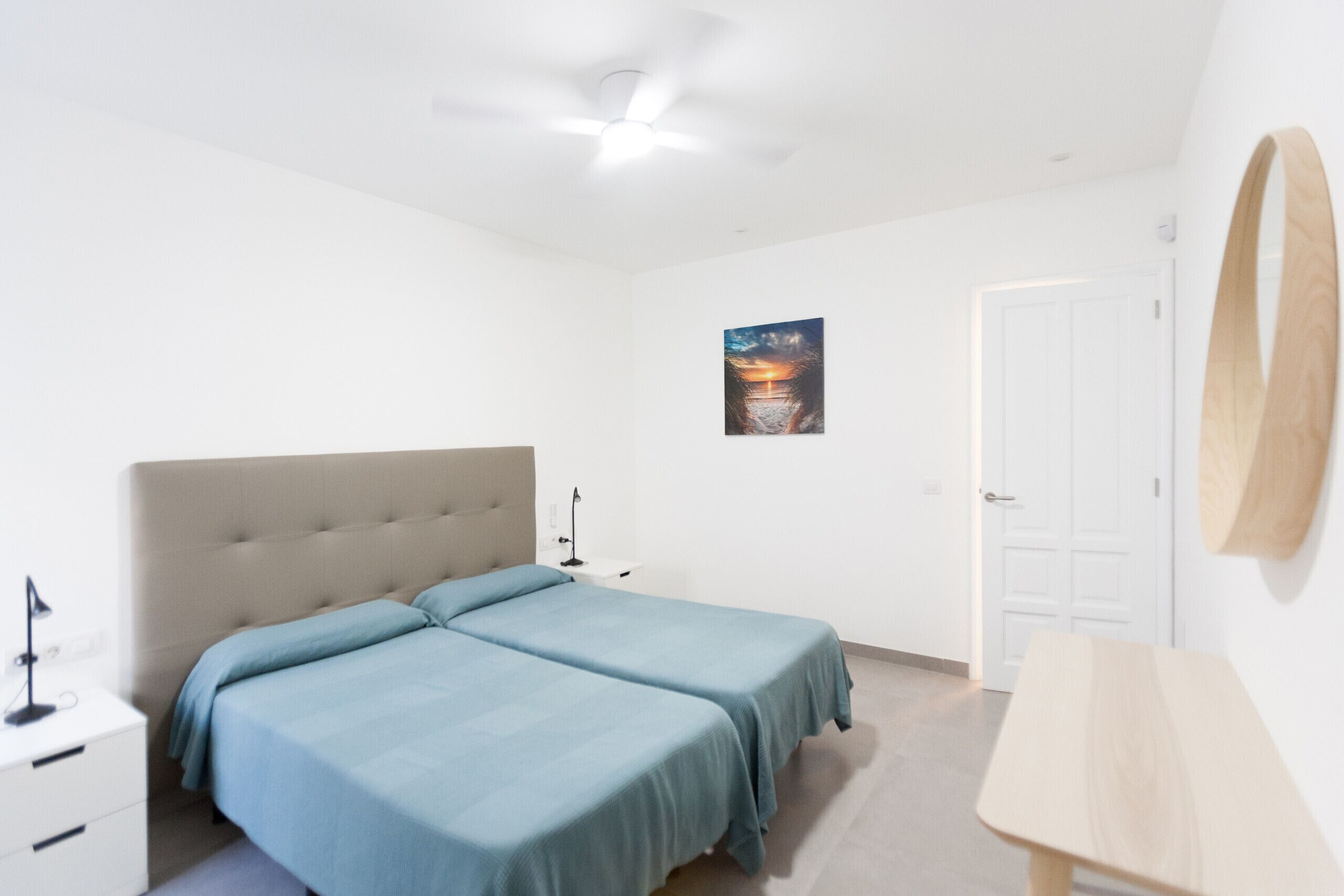 Ett sovrum med två enkelsängar, en takfläkt, ett sängbord med en lampa, en dörr och en spegel som är monterad på väggen.