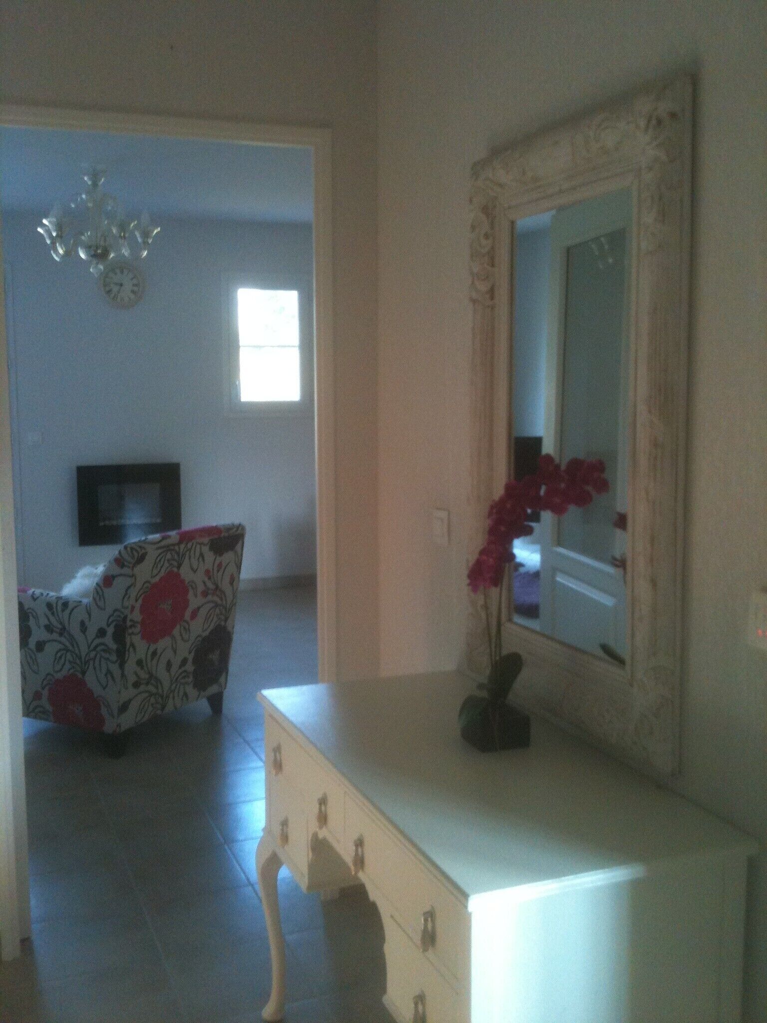 A room with a white dresser, a mirror, and a floral chair. There is a television in the background and a window with natural light.