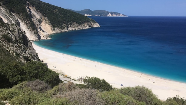 A coastal beach with white sand, clear blue water, and rocky cliffs.
