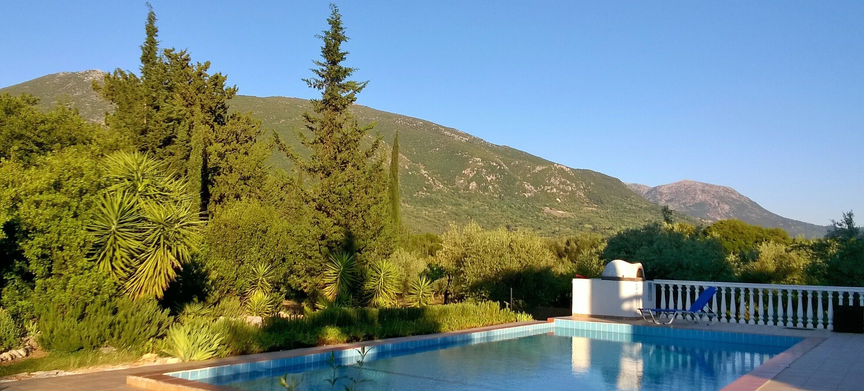 A pool with a mountain backdrop, surrounded by lush greenery and a white building with a blue chair.