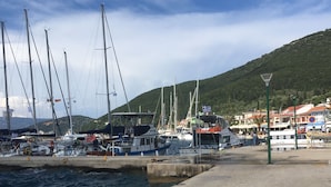A marina with multiple sailboats docked, a small boat with a Greek flag, and a backdrop of a hilly landscape.