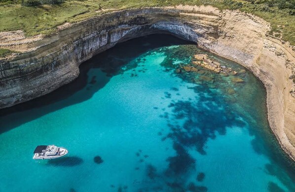 Vista aérea de una zona costera con una gran formación natural en arco, aguas turquesas cristalinas y un pequeño bote anclado cerca del arco.