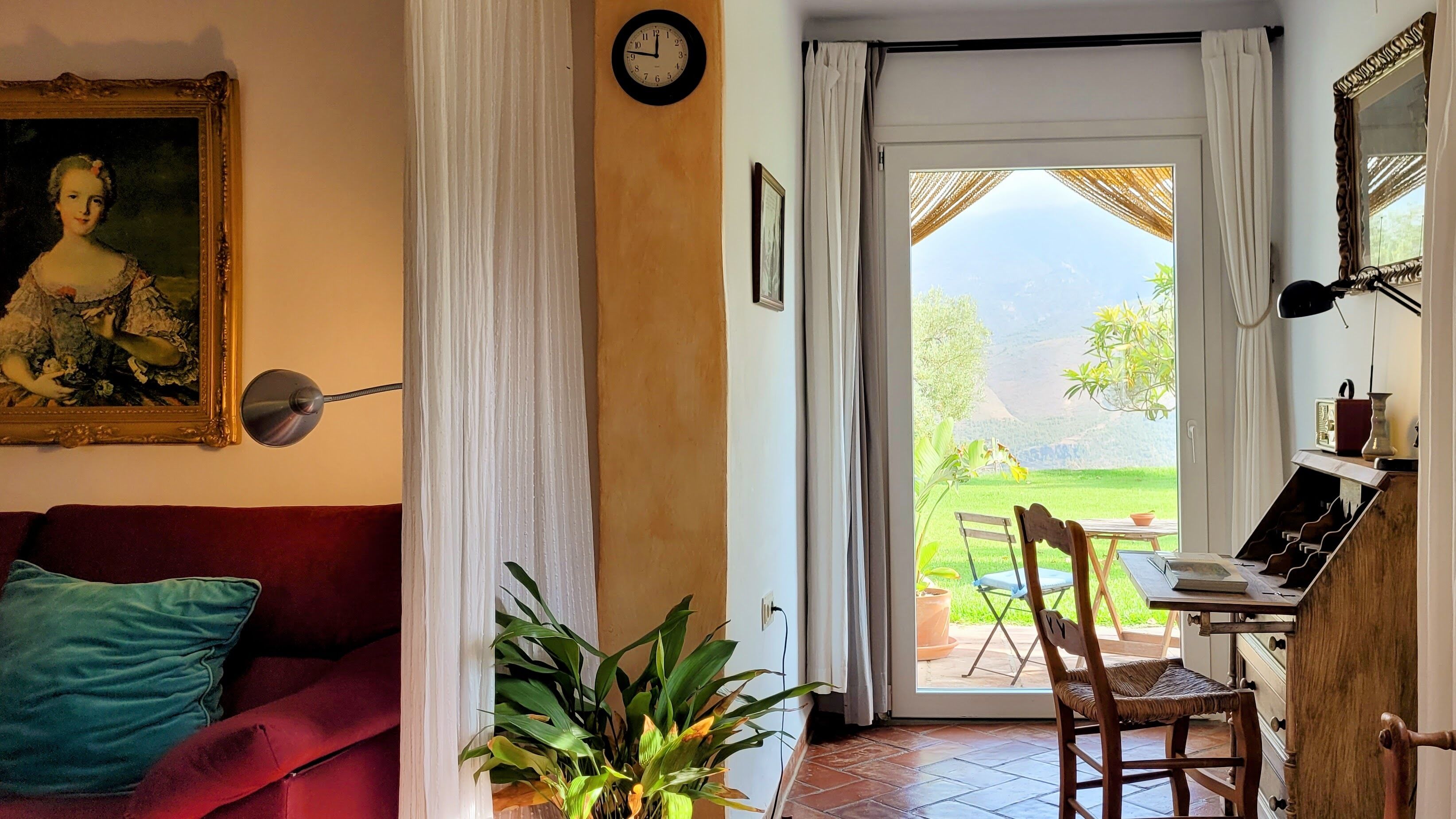 A room with a red sofa, a potted plant, a wooden chair, a table with books, and a view of a garden through a glass door.