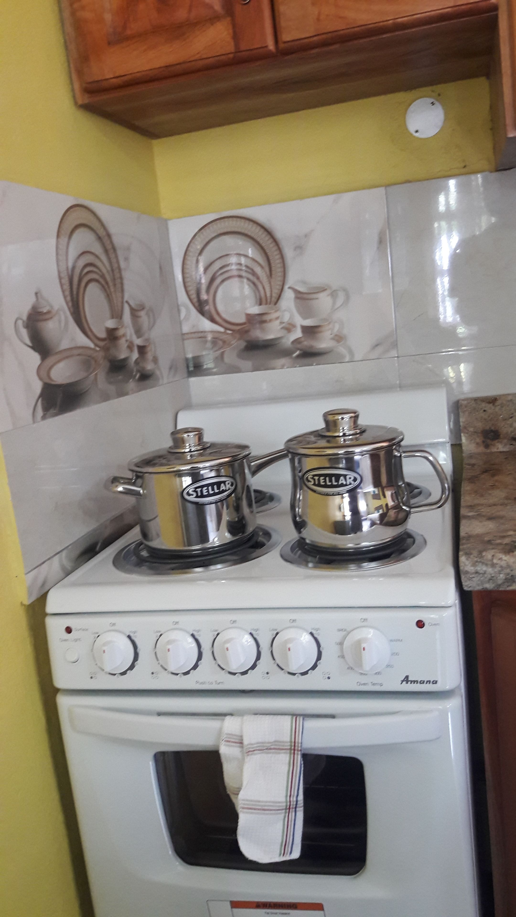 Une cuisine avec un fourneau blanc, deux casseroles en acier inoxydable portant l’inscription « Stellar », et un pare-feu en carreaux orné de motifs d’ustensiles de cuisine.