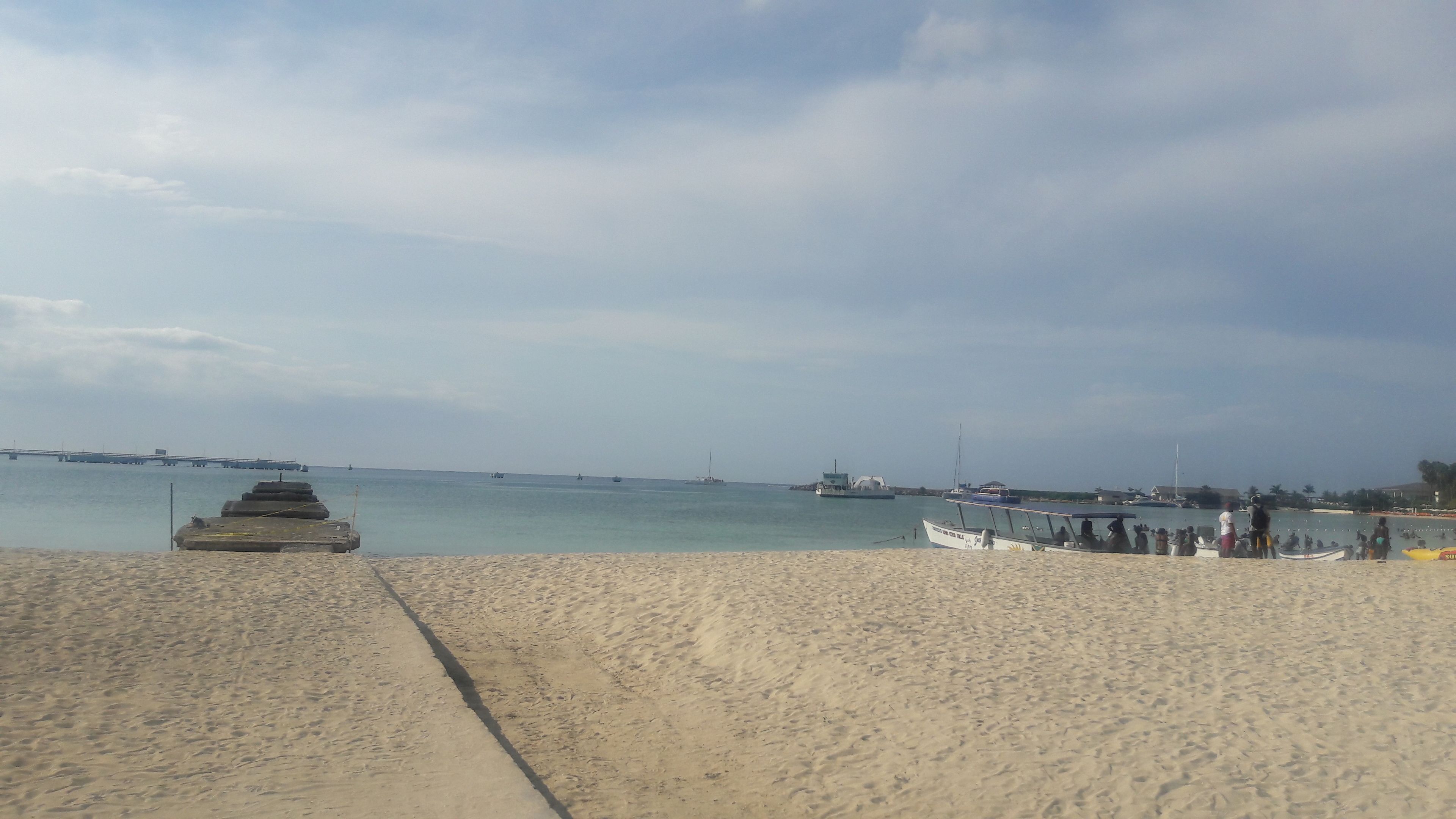 Una playa con un paseo de concreto, barcos y un puente a lo lejos.