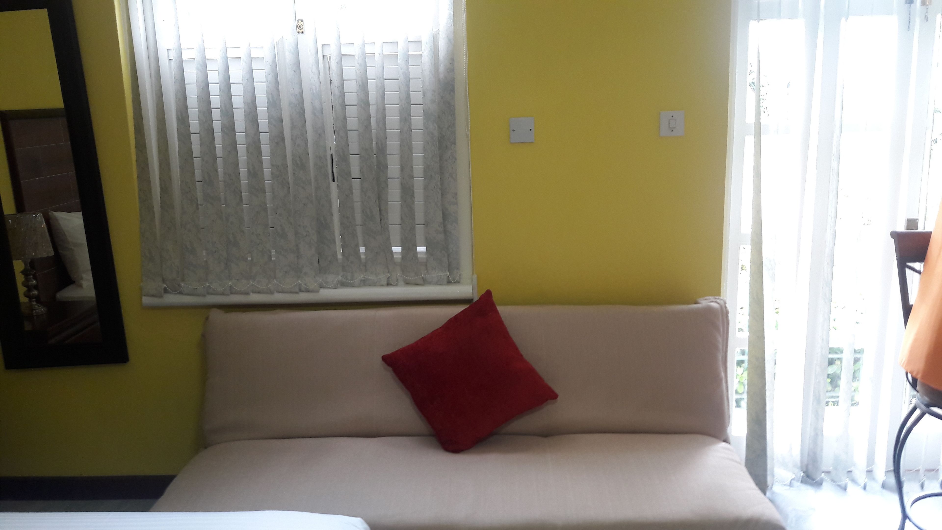 Une pièce avec un canapé beige, un coussin rouge, une fenêtre avec des stores et un miroir au mur.