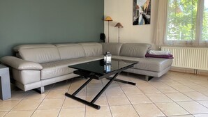 Un salon comprenant un canapé d’angle, une table basse en verre, un lampadaire, un radiateur et une fenêtre avec des rideaux.