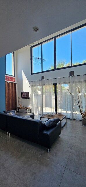 Uma sala de estar ampla, com um sofá preto grande, uma mesa de centro de madeira e uma televisão na parede.