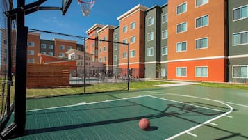 Una cancha de baloncesto al aire libre contigua a un edificio de varios pisos con techo verde y fachada de ladrillo rojo.