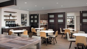 A modern dining area with round tables and chairs, a coffee station with multiple coffee makers, and wooden cabinets with frosted glass panels.