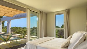 A bedroom with a large bed, a balcony with chairs, and a view of greenery.