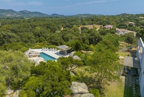 Zona de piscina con sillones de descanso y un edificio pequeño, rodeada de abundante vegetación y colinas al fondo.