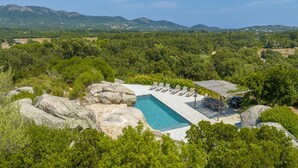 A rectangular pool with a stone deck, surrounded by large rocks and lush greenery, with mountains in the background.