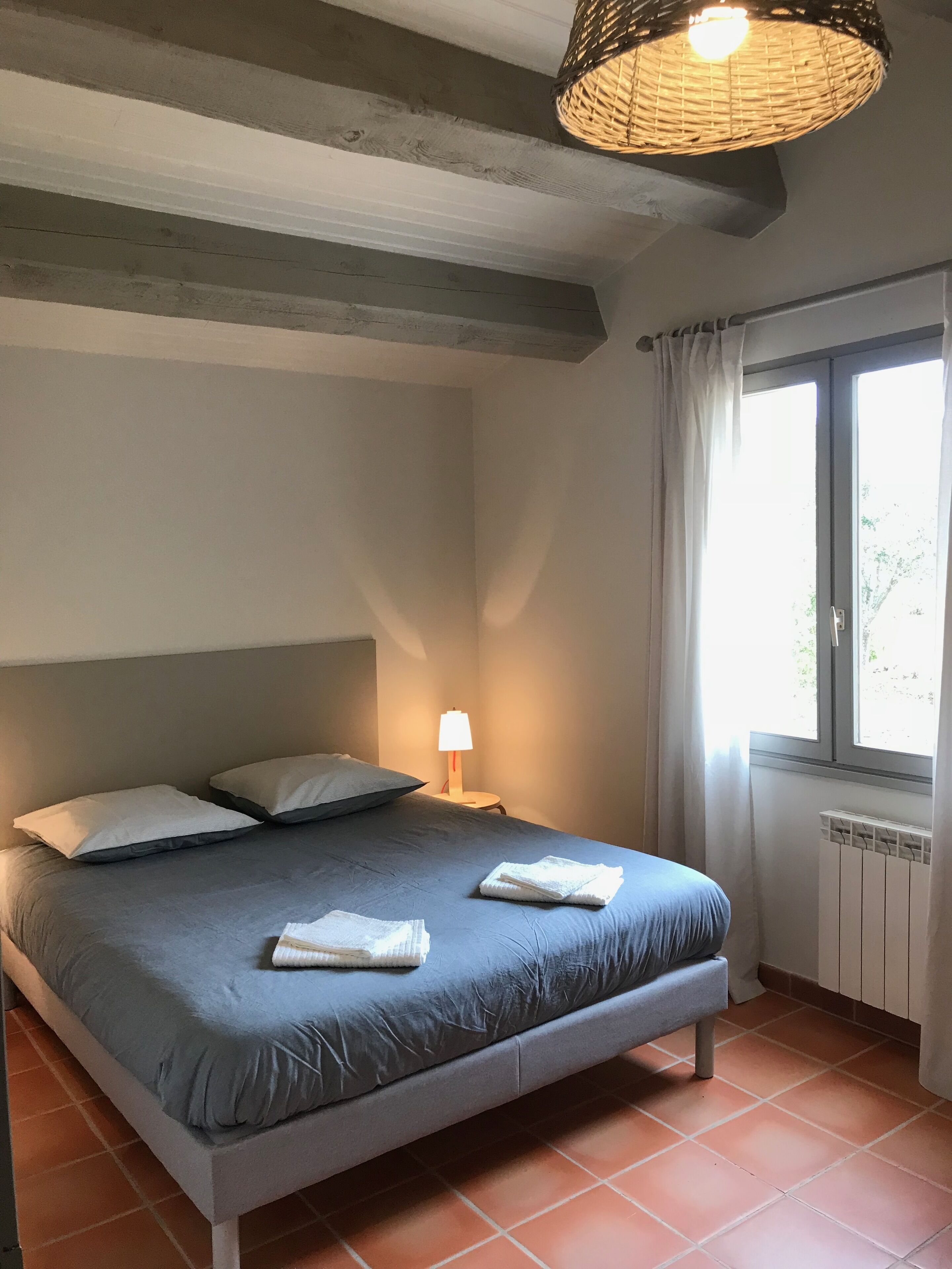 Un dormitorio con cama, almohadas, lámpara de noche, ventana con cortinas y radiador.
