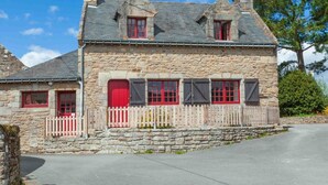Kivestä rakennettu talo, jossa on harjakatto, punaiset ikkunaluukut ja koristeellinen aita.
