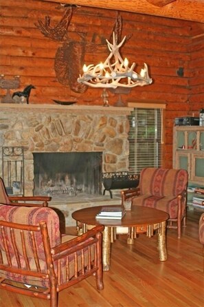Cozy cabin interior with a stone fireplace, wooden ceiling, and chandelier with antler motifs.