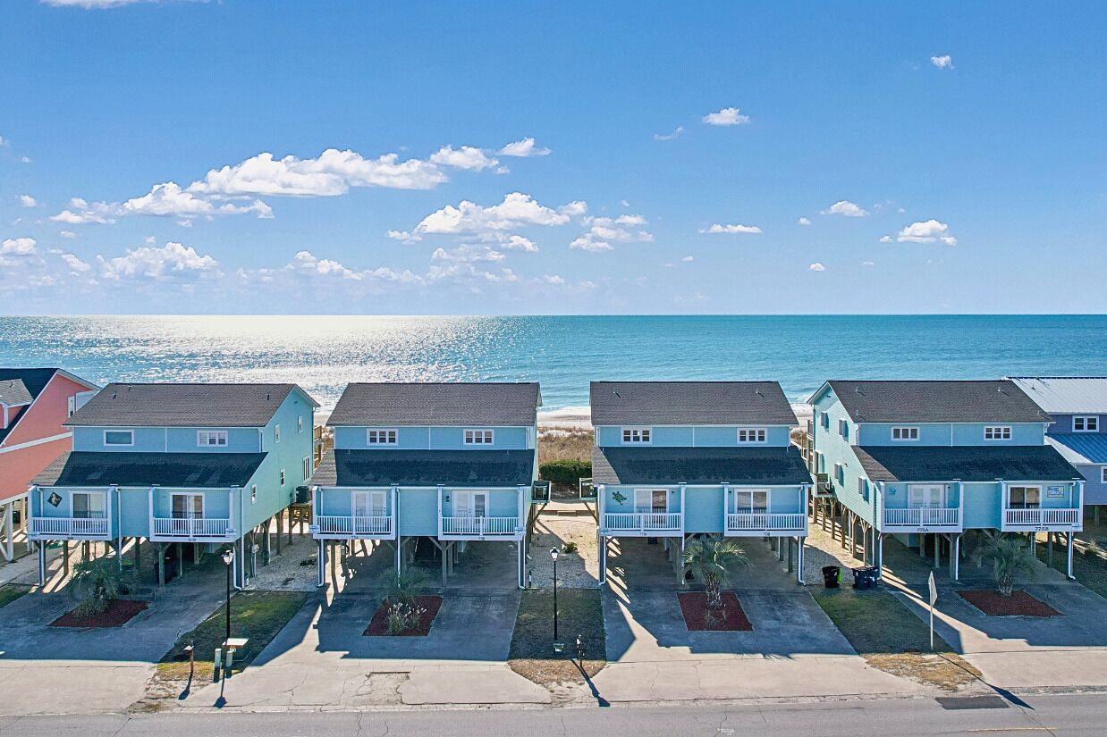 Una hilera de casas frente a la playa con balcones, cielo despejado y el océano al fondo.