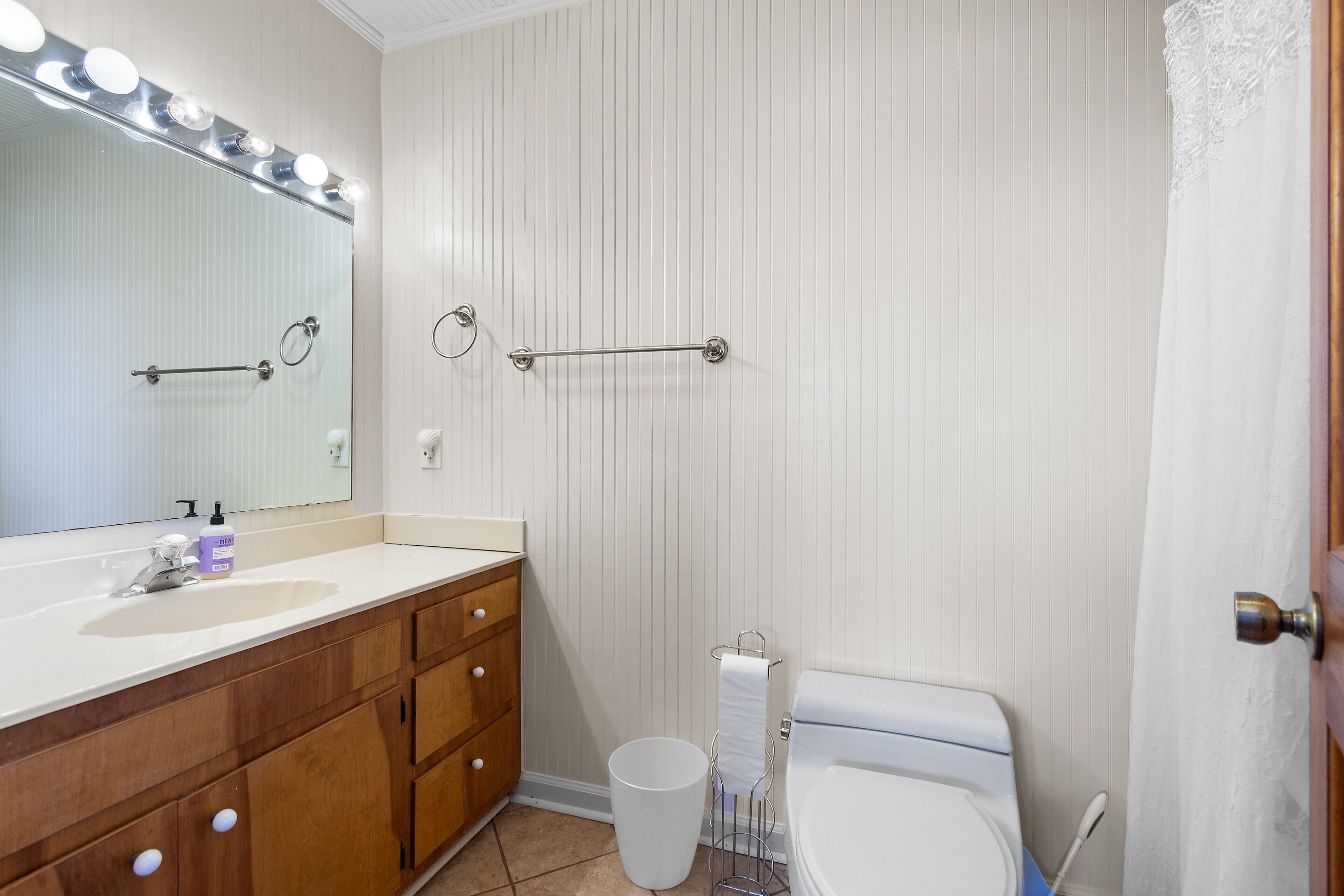 A bathroom with a vanity, mirror, lights, towel rack, toilet, and trash can.