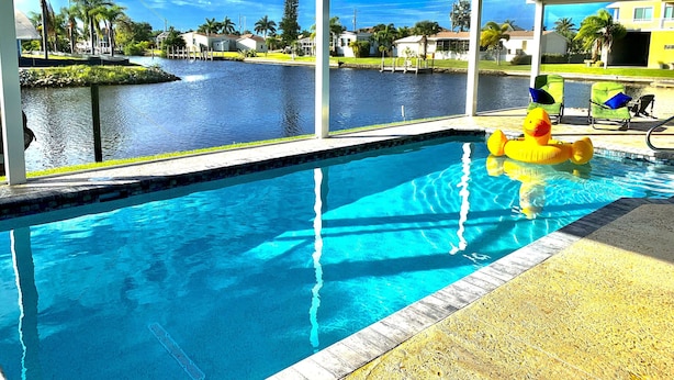 Una piscina con agua cristalina azul, un patito inflable amarillo y vistas a una zona residencial con palmeras y casas.