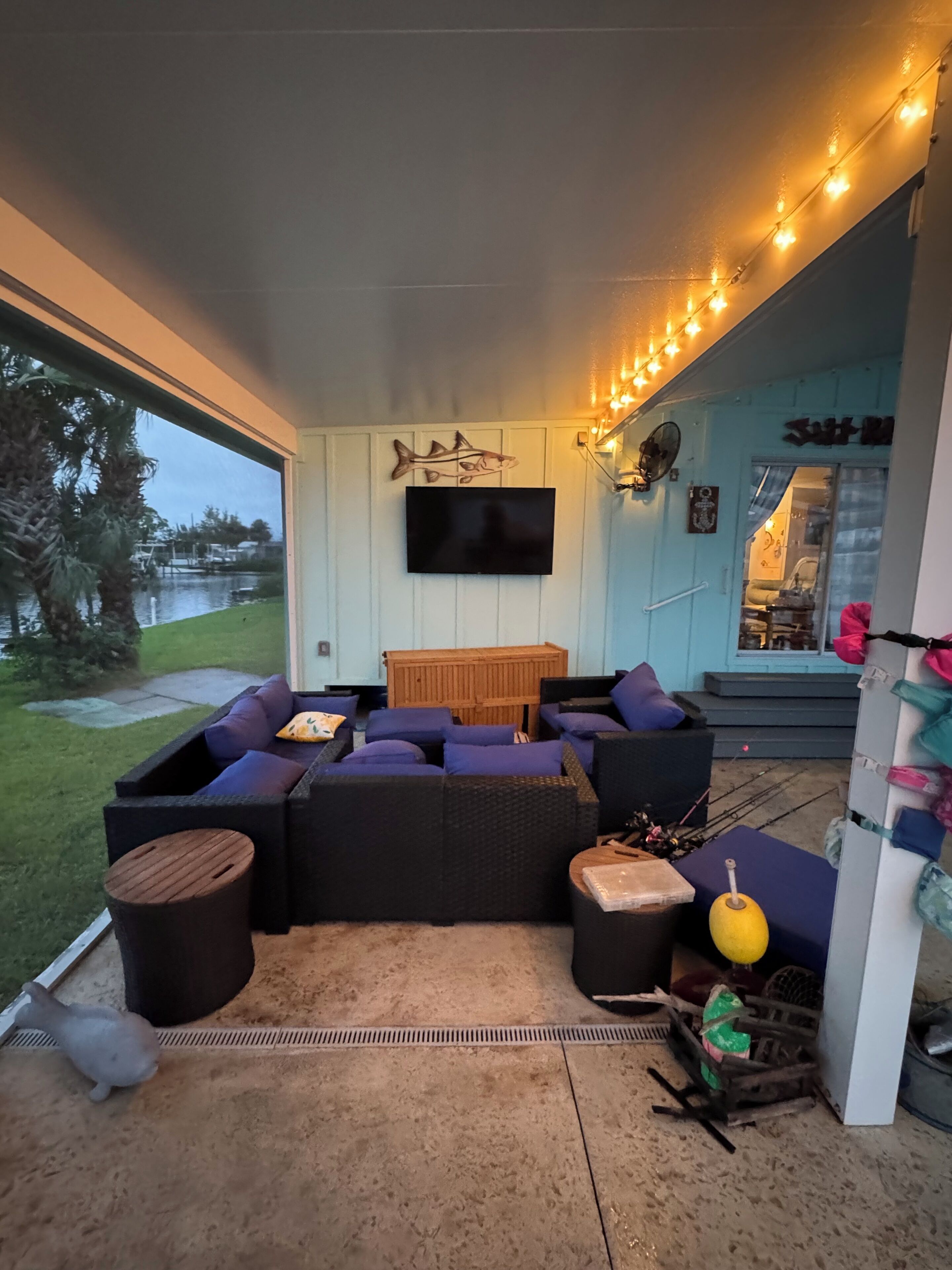 A covered outdoor seating area with a television mounted on the wall, a wooden fish decoration, and string lights.