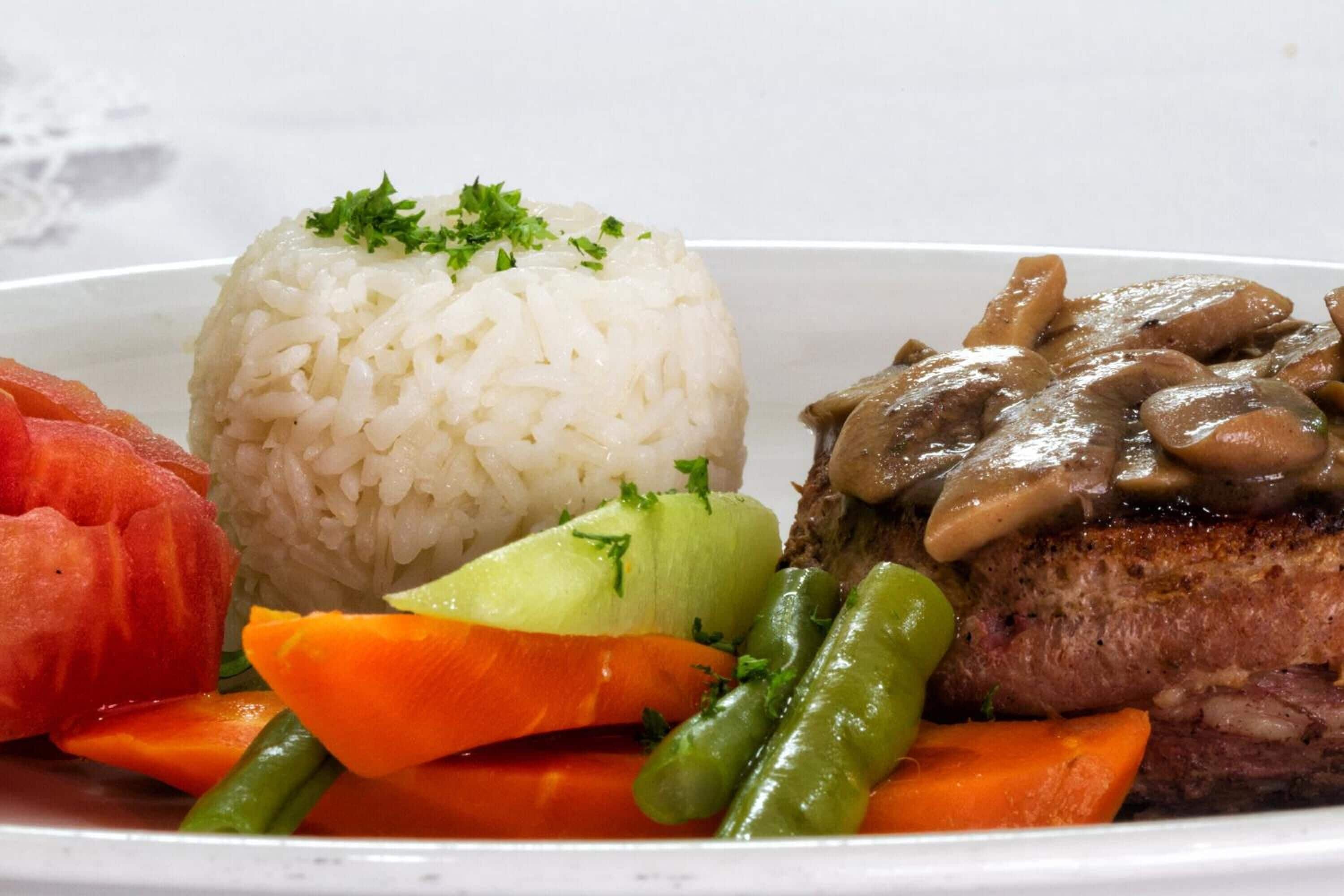 Un piatto con riso bianco, una bistecca di manzo condita con funghi e verdure miste, tra cui carote e fagiolini.