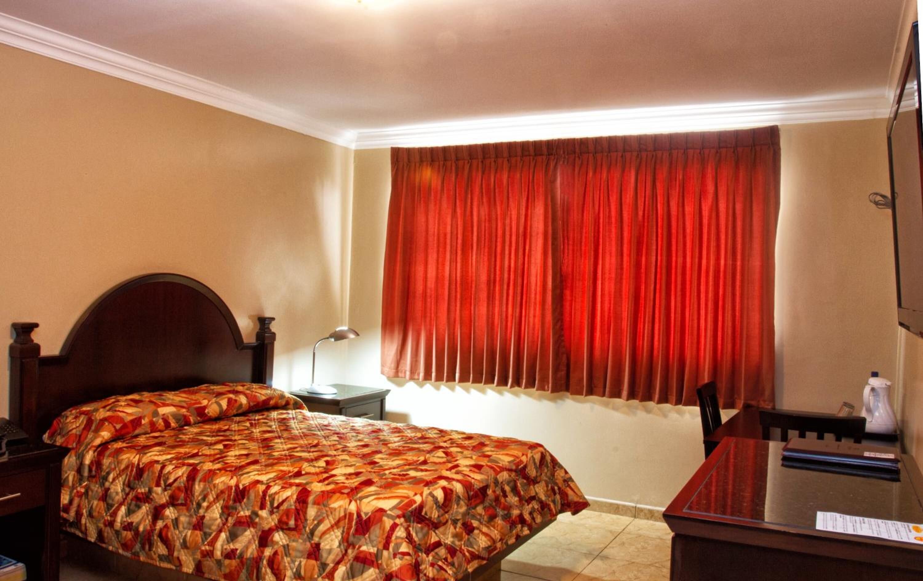 A hotel room with a bed, a desk, a chair, a lamp, and a window with red curtains.