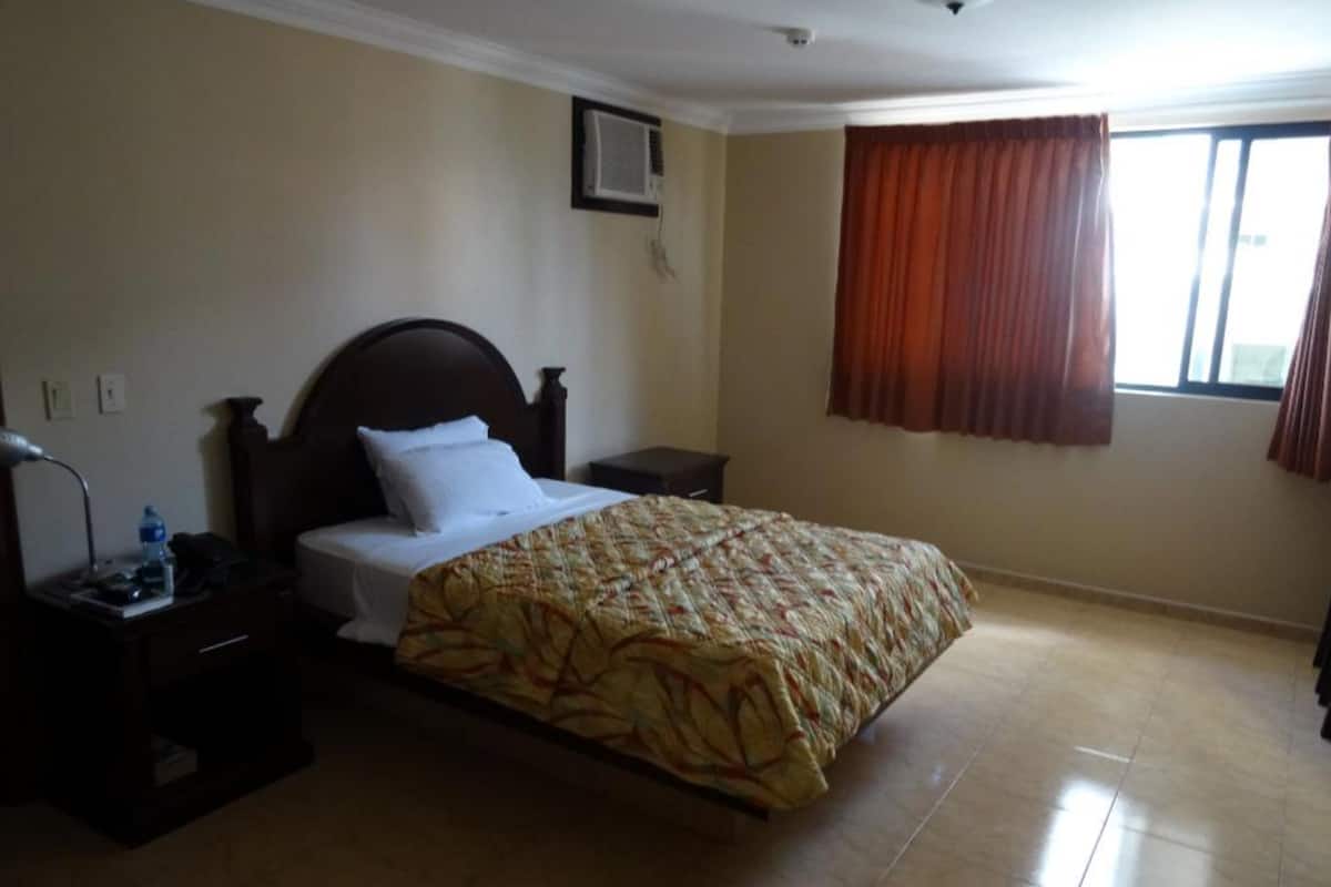 A hotel room with a bed, a nightstand, a lamp, a water bottle, and a window with curtains.