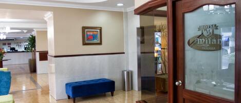 A hotel lobby with a reception desk, a blue bench, a trash can, and a framed picture on the wall.