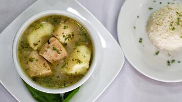 Un tazón de sopa con trozos de carne y papas, adornada con hierbas, acompañado de arroz.