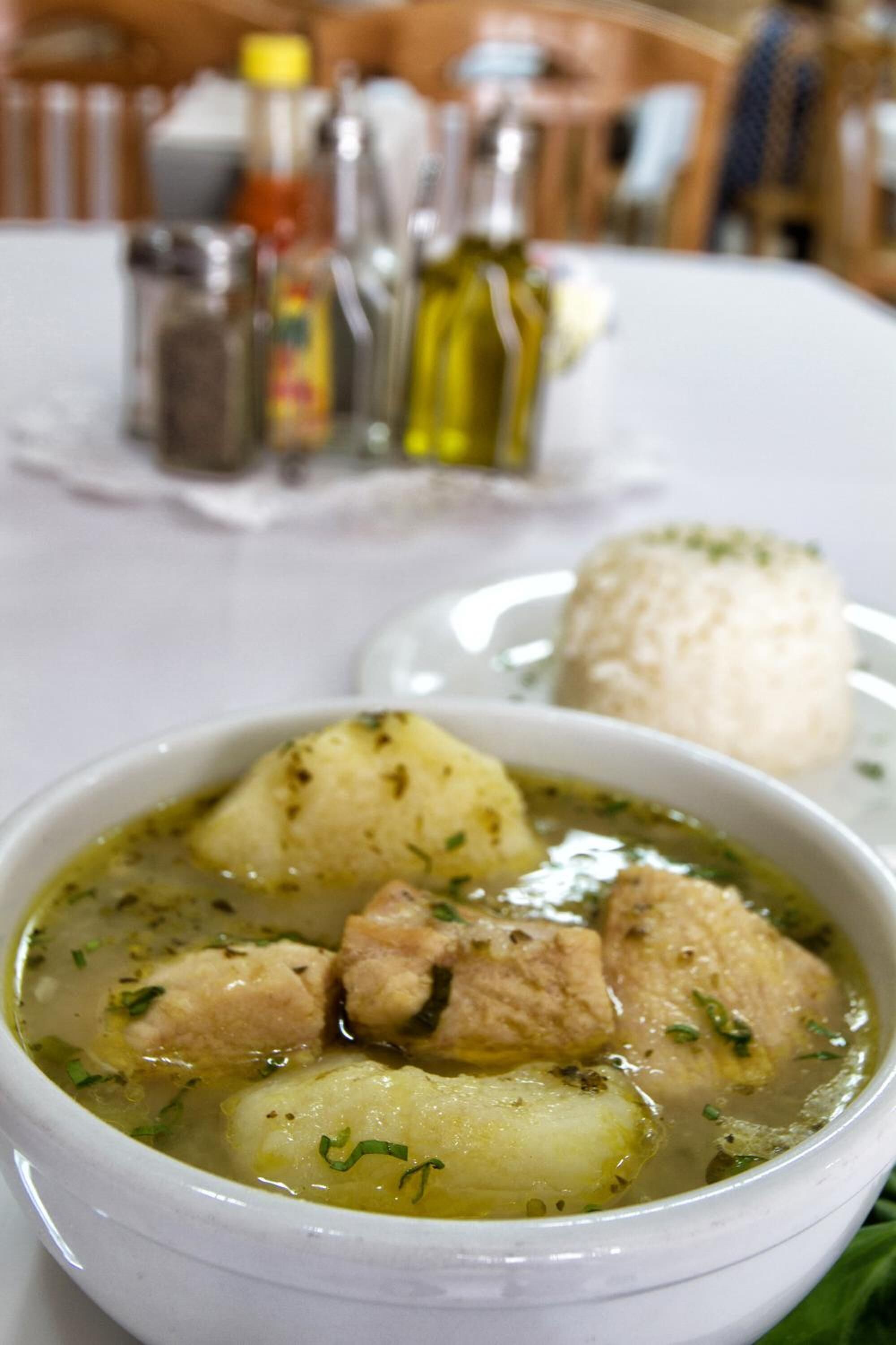 Uma tigela de sopa com batatas e carne, um prato branco com um monte de arroz e uma mesa com saleiros e pimenteiros ao fundo.