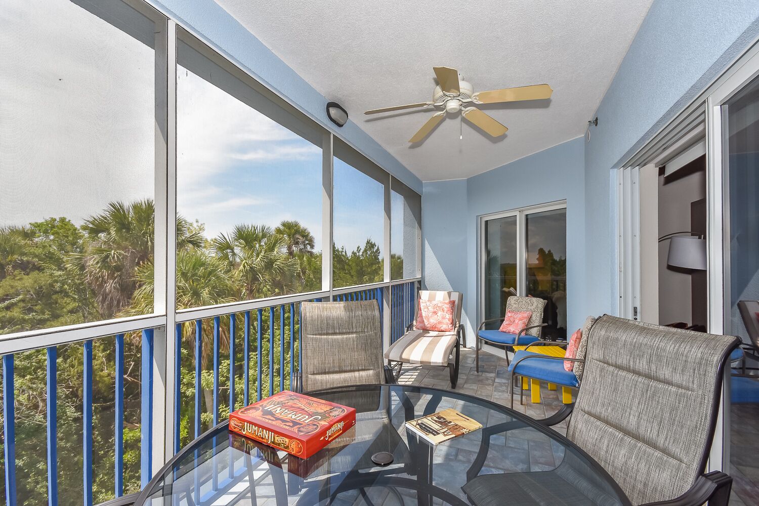 Un balcon avec une table en verre, deux chaises, un ventilateur de plafond et une vue sur des palmiers.