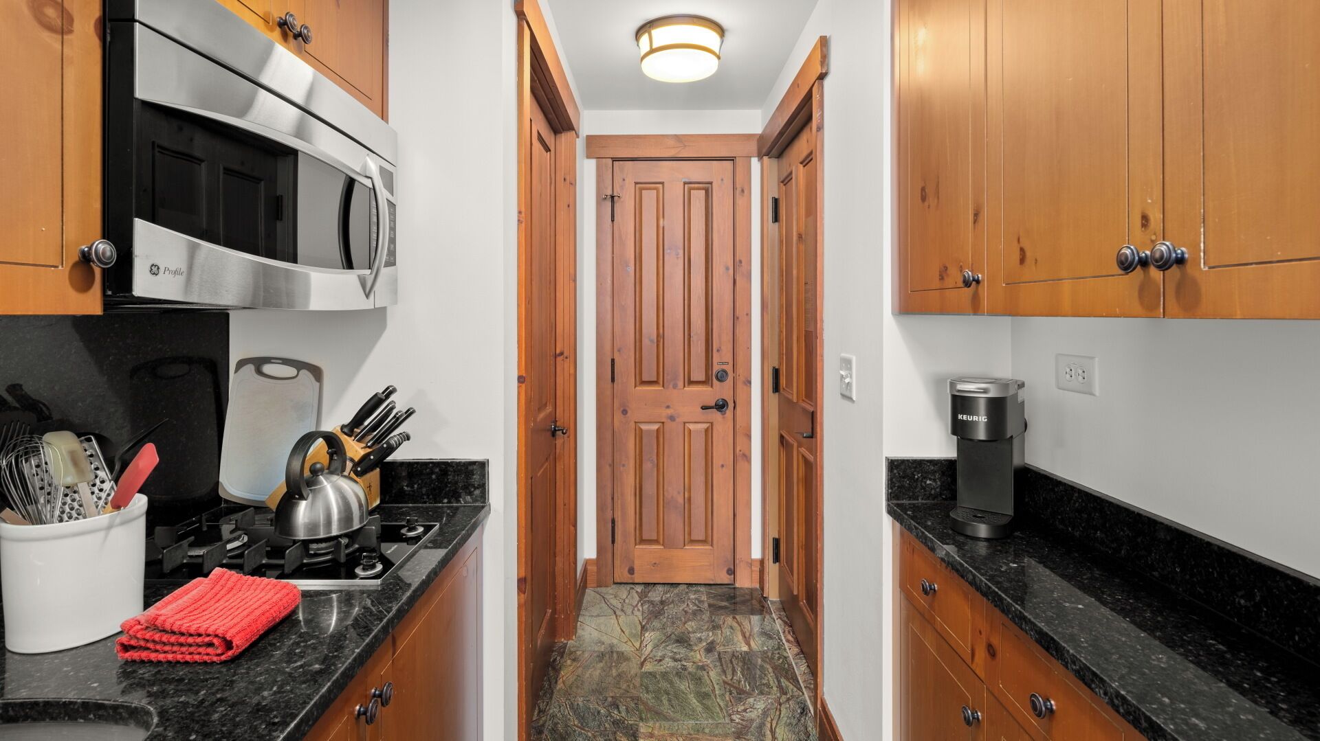 A narrow kitchen with wooden cabinets, a stainless steel microwave, a kettle, and a Keurig coffee maker.