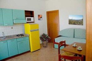 A compact kitchen with teal cabinets, a yellow refrigerator, a microwave, and a dining area with a wooden table and chairs. - Luzia Agriresort (Ricadi)