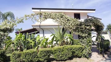 Un bâtiment à deux étages, doté d’une façade blanche et d’un toit en tuiles, entouré d’une végétation luxuriante et de plantes en fleurs.