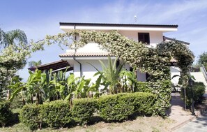 A two-story building with a white facade and a tiled roof, surrounded by lush greenery and flowering plants. - Luzia Agriresort (Ricadi)