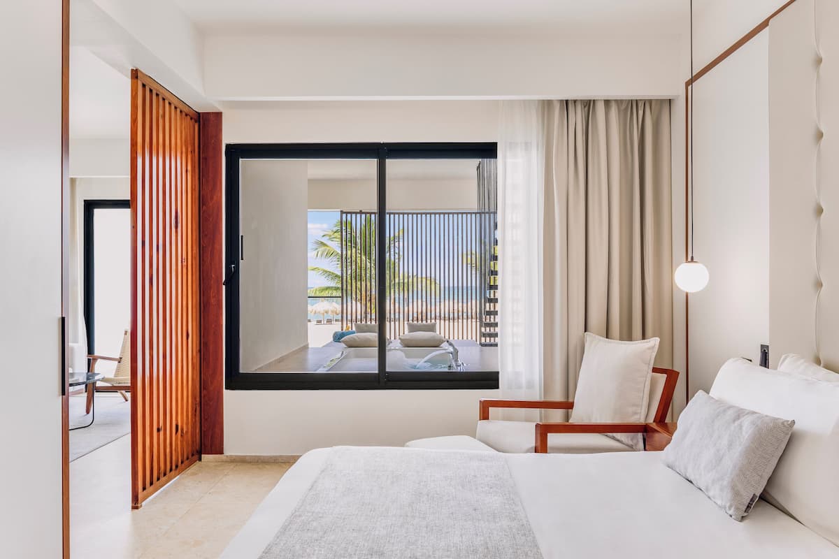 Ein Hotelzimmer mit Bett, Sessel, Blick auf Pool und Palmen sowie einem Balkon mit Vorhängen.