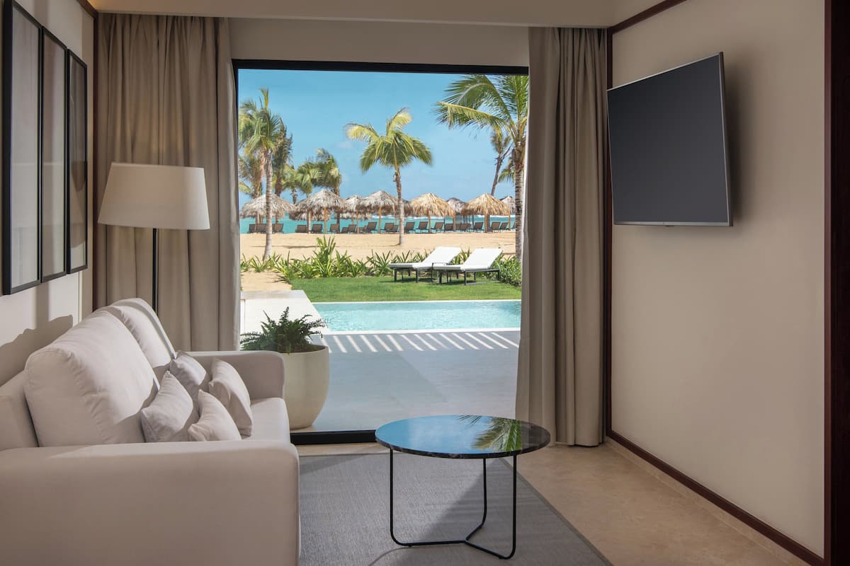 Ein Hotelzimmer mit Sofa, einem kleinen Tisch, einem Fernseher und Blick auf einen Pool sowie einen Strand mit Liegestühlen.