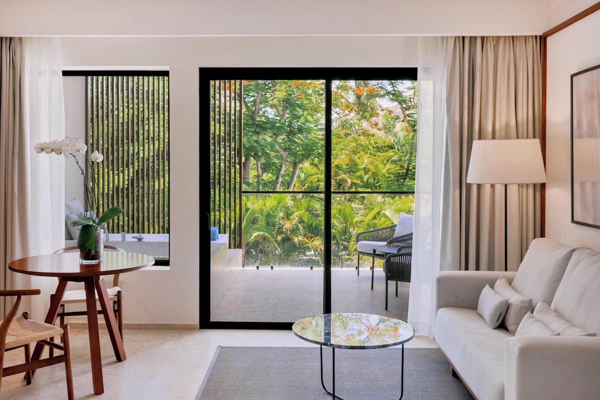 Ein modernes Wohnzimmer mit einer Couch, einem kleinen runden Tisch, einem Essbereich mit Tisch und Stühlen sowie einer großen Schiebeglasdoor zum Außenbalkon mit Pflanzen und Blick auf Bäume.