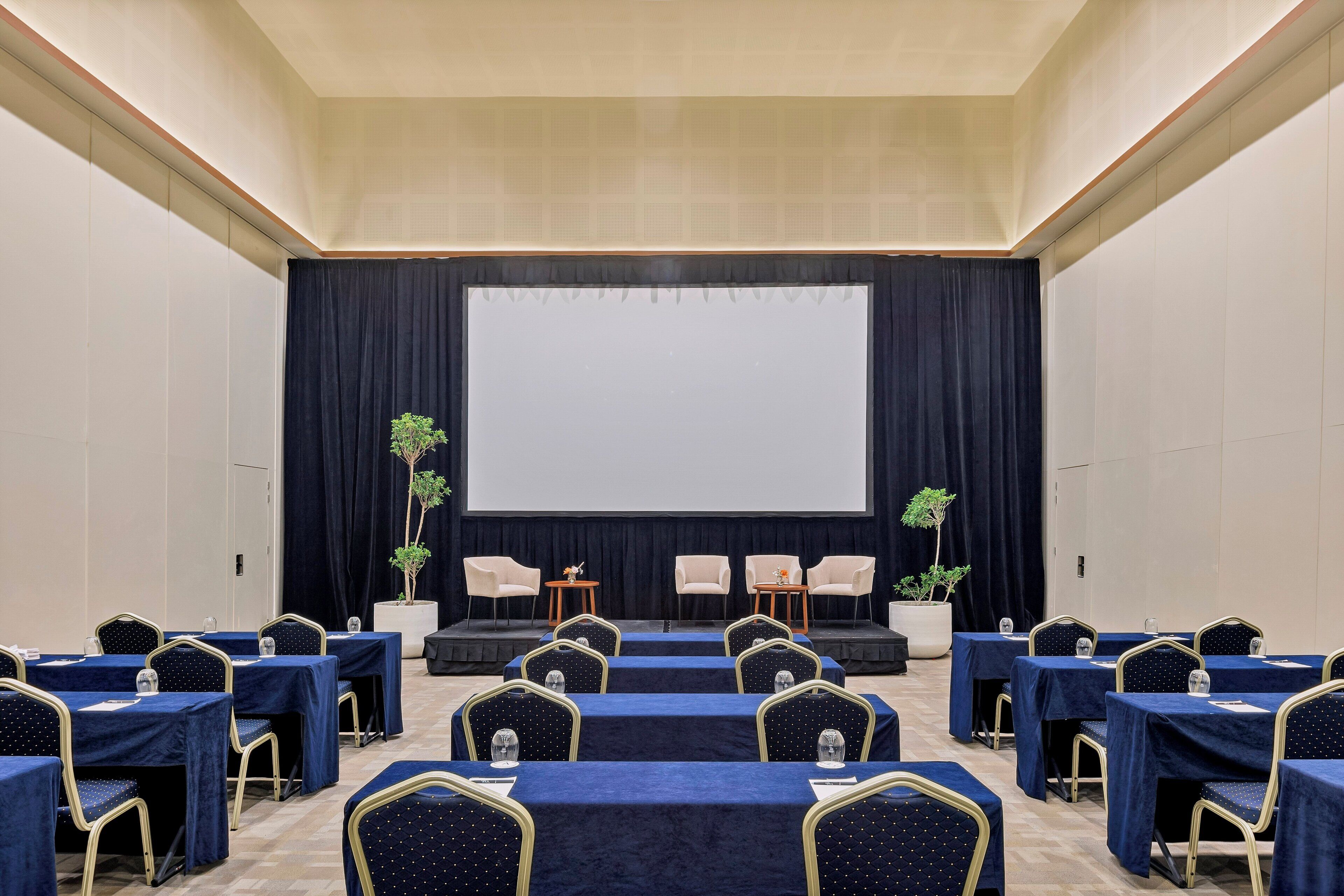 A conference room with a stage, rows of tables with chairs, and potted plants.
