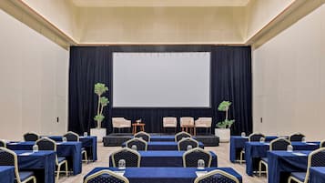A conference room with a stage, rows of tables with chairs, and potted plants.