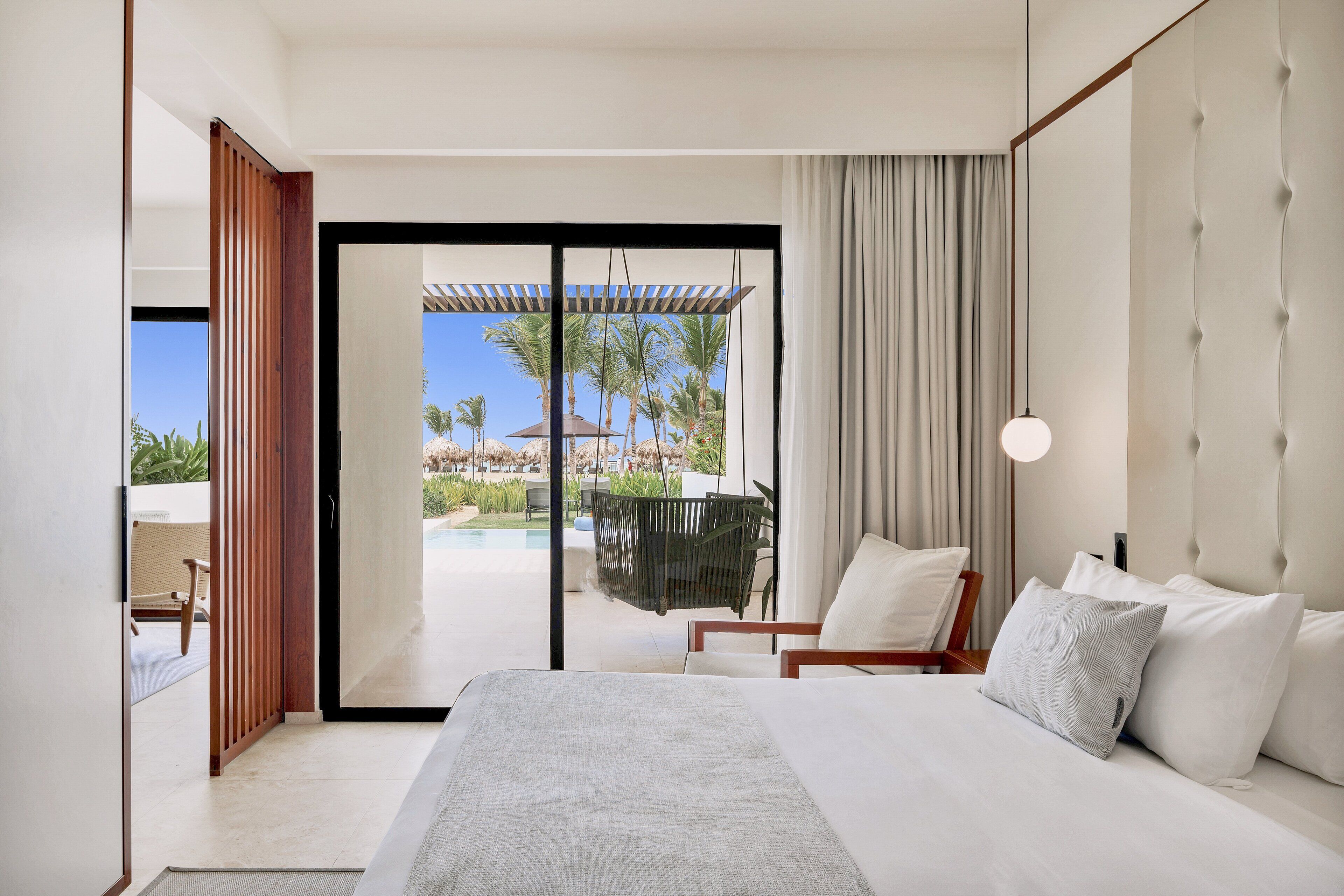 A modern hotel room with a large bed, a view of a pool and palm trees, and a sliding glass door leading to an outdoor area.