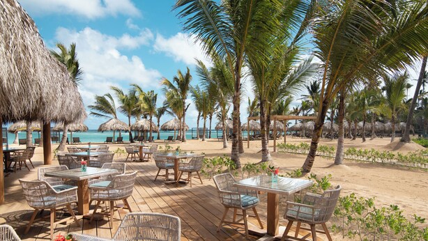Een terras met rieten meubels en rieten parasols, met uitzicht op een strand.
