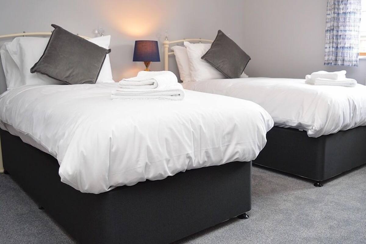 Une chambre d’hôtel avec deux lits, chacun doté de draps blancs et d’une tête de lit sombre. Une lampe se trouve sur la table de chevet entre les deux lits, et une fenêtre avec des rideaux est située à droite.