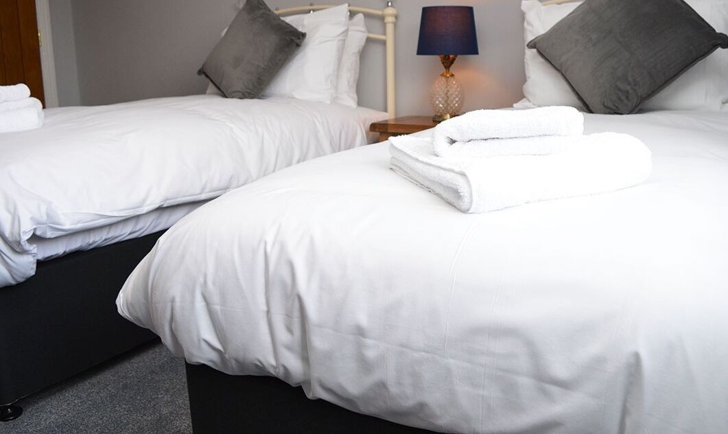 Two white beds with grey pillows, a small bedside table with a lamp, and a folded white towel on the right bed.