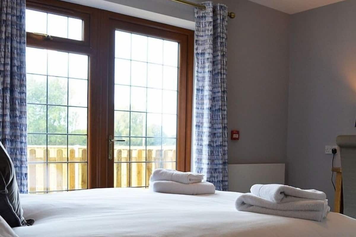 Une chambre d’hôtel avec un grand lit, deux fenêtres avec des rideaux et une vue sur une terrasse en bois à l’extérieur.