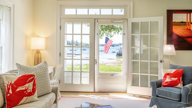 A living room with a view of a lake, a door with glass panels, and a lamp on a side table. - Dew Drop 2 Village Cottage, Steps to Harbor, Lawn & Boathouse (Port Clyde)