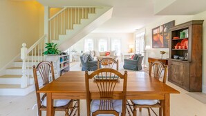 A dining area with a wooden table and chairs, a staircase, and a kitchen area with cabinets and a dining table. - Dew Drop 2 Village Cottage, Steps to Harbor, Lawn & Boathouse (Port Clyde)