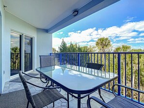 A balcony with a glass table and chairs, overlooking a tropical landscape with trees and a clear sky.