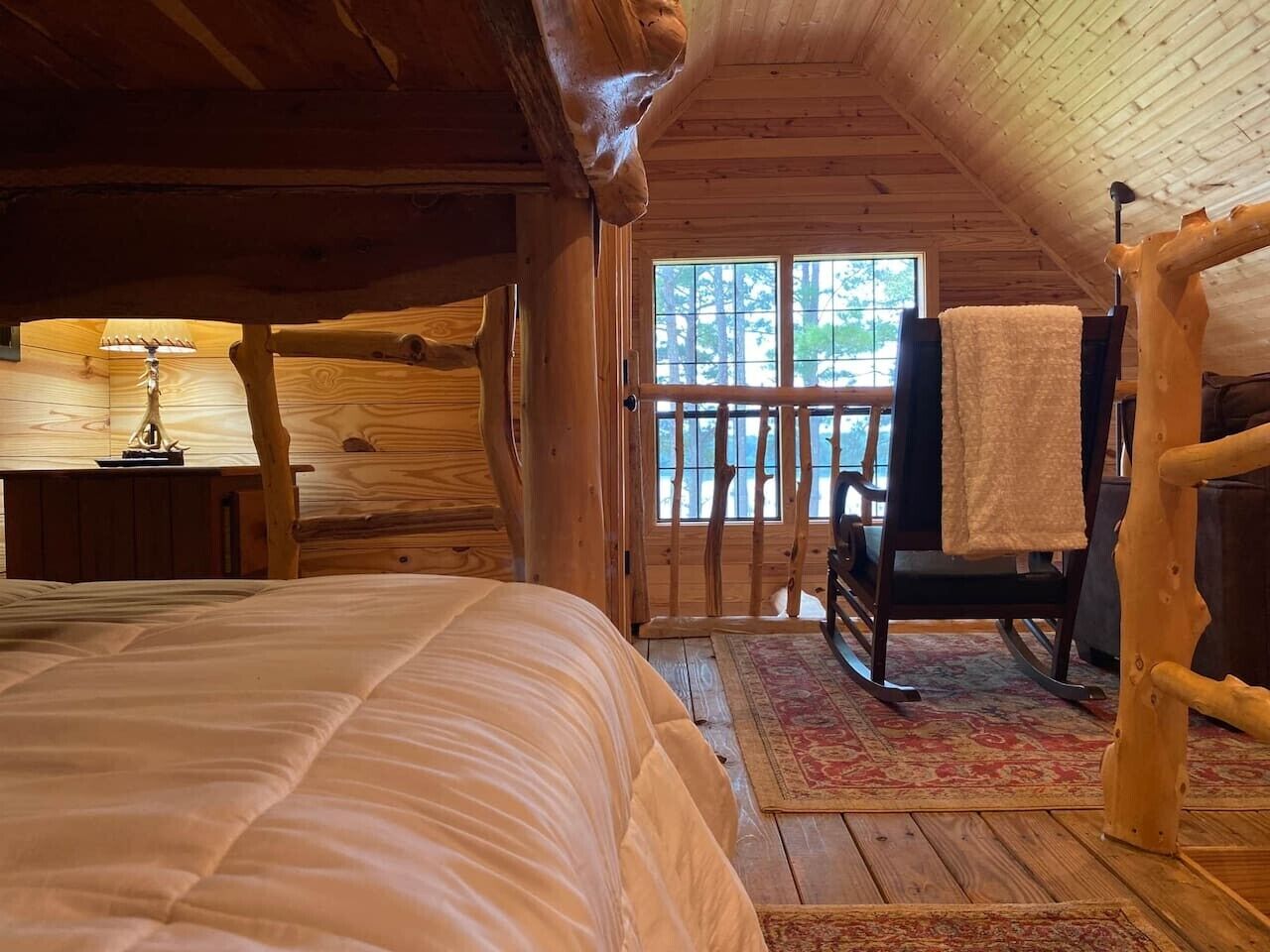 Une chambre à coucher située dans un chalet en rondins, avec un lit, un fauteuil à bascule, une lampe et une vue sur l’extérieur.